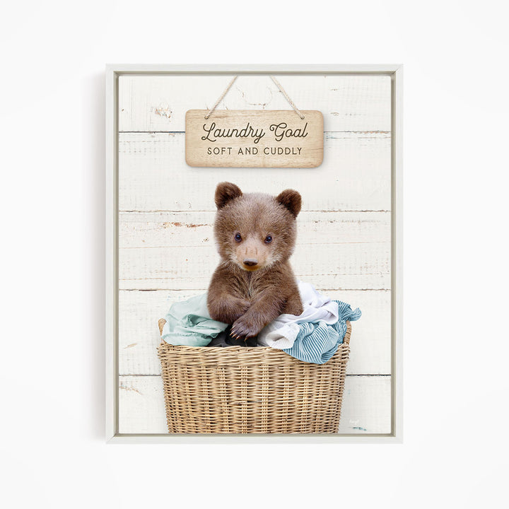 A cute brown bear sitting in a wicker laundry basket, with a wooden sign above that says "Laundry Goal Soft and Cuddly".