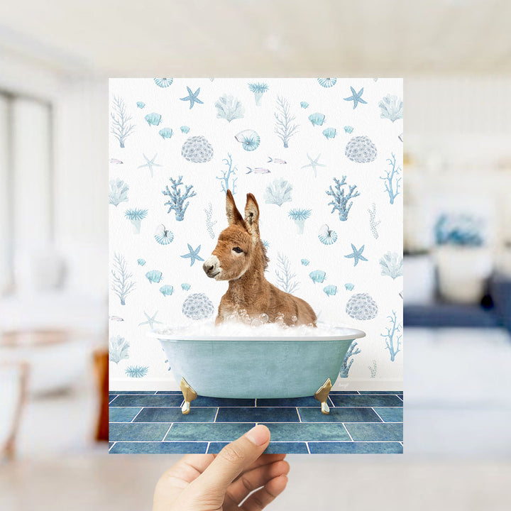 A hand is holding up a framed image of a donkey sitting in a bathtub filled with bubbles, surrounded by a pattern of seashells, starfish, and other marine life.