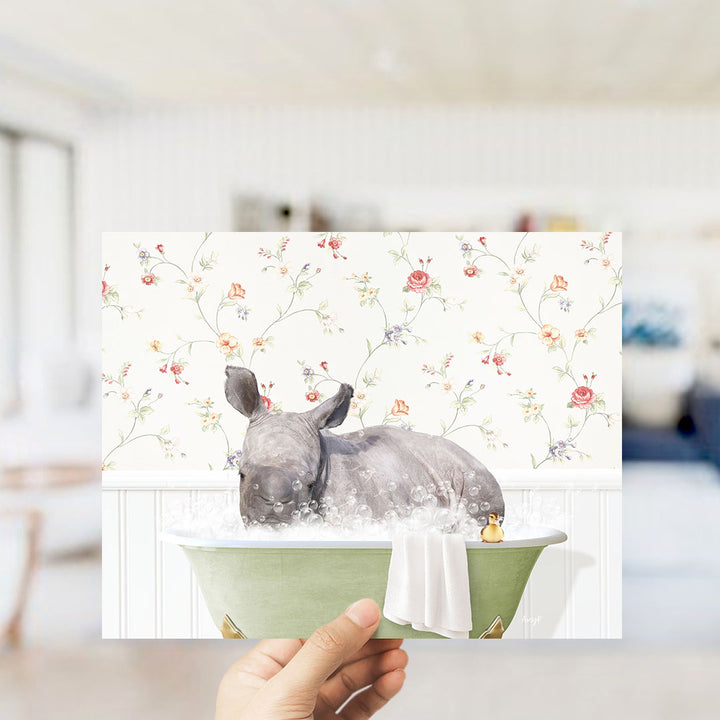A hand holding a framed image of a rhinoceros sitting in a bathtub filled with bubbles, with a floral wallpaper in the background.