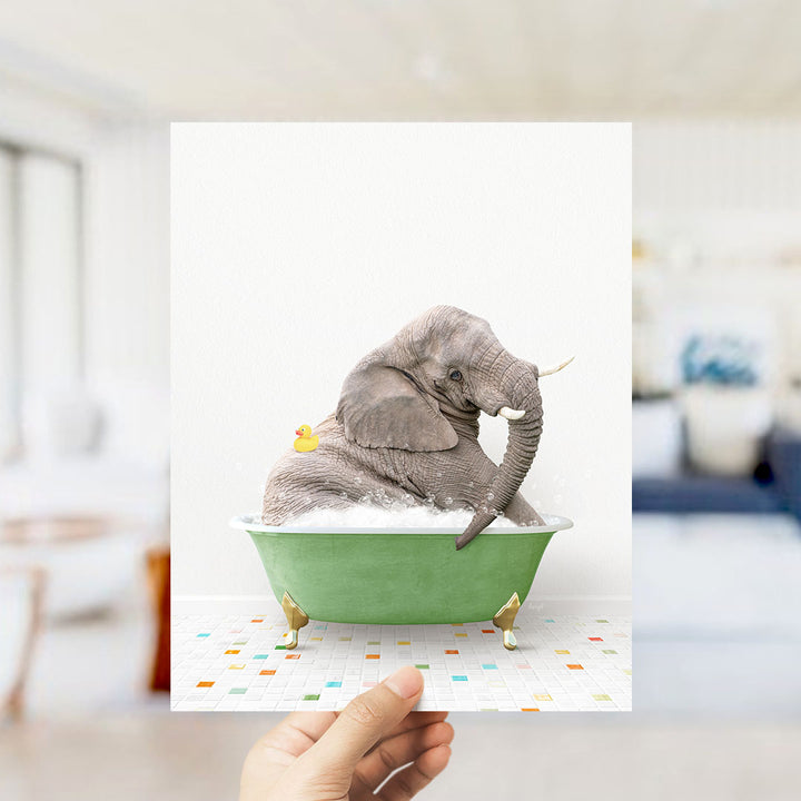A hand holds up a framed image of a gray elephant sitting in a green bathtub, with a yellow rubber duck floating nearby.