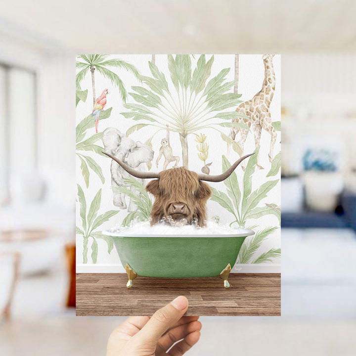 A hand holds up a framed print featuring an illustration of a horned animal, likely a yak or a yak, sitting in a green bathtub surrounded by tropical foliage and other animals.