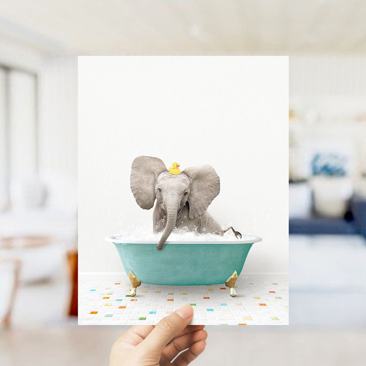 A hand holds up a framed image of a baby elephant sitting in a bathtub filled with bubbles, wearing a yellow rubber duck.