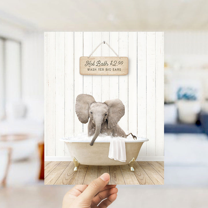 A hand holding a framed image of an elephant sitting in a bathtub, with a sign above it advertising "Hot Bath $2.00 WASH EARS".