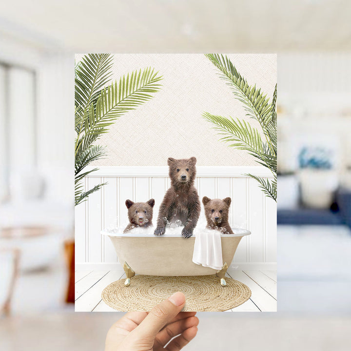 A hand holds up a framed image depicting three adorable brown bears sitting in a bathtub, surrounded by lush green palm leaves.