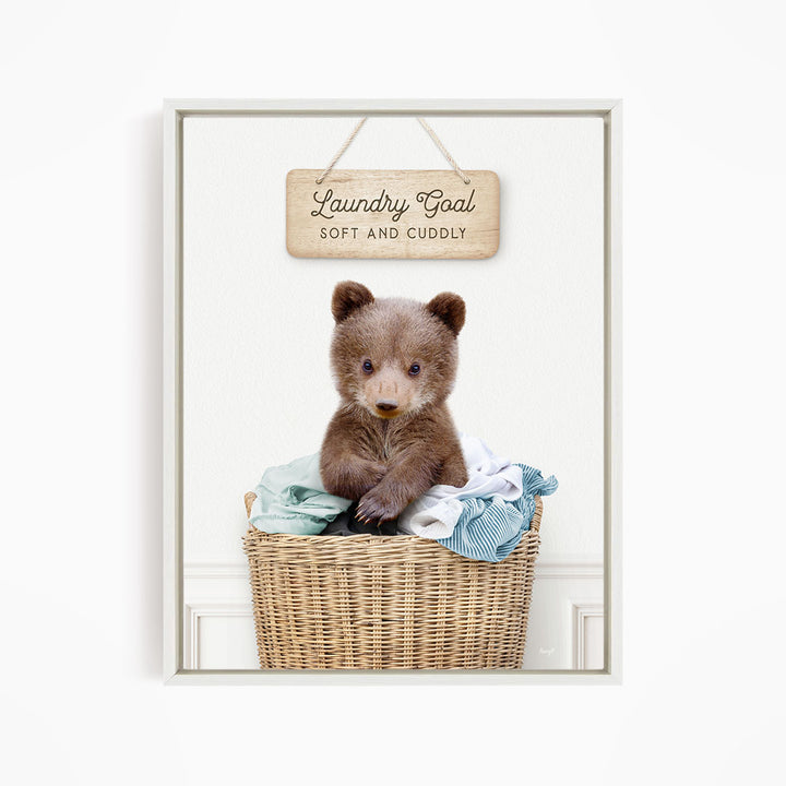 A cute brown bear sitting in a wicker laundry basket, with a wooden sign above that says "Laundry Goal Soft and Cuddly".