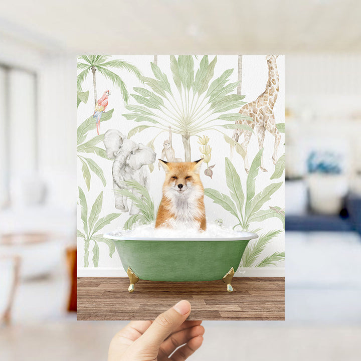 A hand holds up a framed print featuring a fox sitting in a bathtub surrounded by tropical plants and animals, including a giraffe and a parrot.