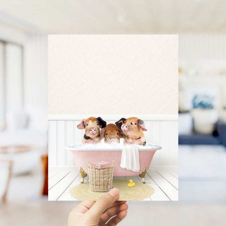 A hand holds up a framed image depicting three adorable pigs sitting in a pink bathtub, surrounded by a yellow rubber duck and a white towel.