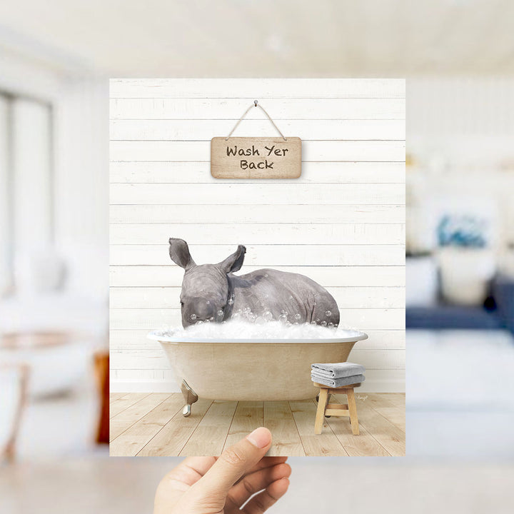 A hand holds up a framed image depicting a rhinoceros sitting in a bathtub filled with water, with a sign above that reads "Wash Yer Back".