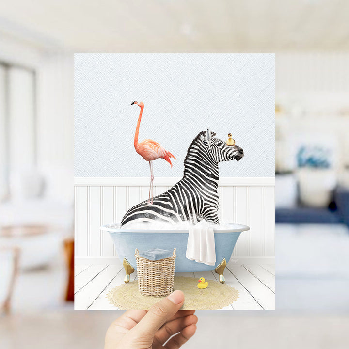 A hand holds up a framed image depicting a zebra sitting in a bathtub, with a pink flamingo standing on its back and a yellow rubber duck floating nearby.