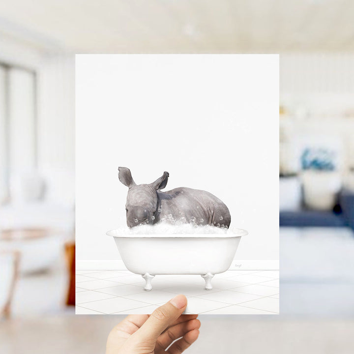 A hand holds up a framed image depicting a rhinoceros sitting in a bathtub filled with water.
