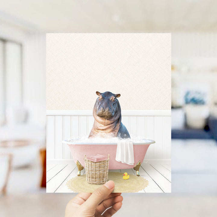 A hand holds up a framed image depicting a hippopotamus sitting in a pink bathtub filled with bubbles, surrounded by a yellow rubber duck and a white towel.