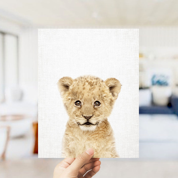 A hand is holding up a square piece of paper with a close-up image of a young lion cub on it.