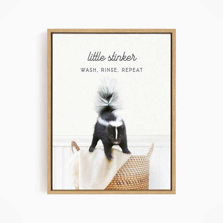 A framed image of a skunk sitting on a woven basket, with the text "little stinker" and "WASH, RINSE, REPEAT" written above it.