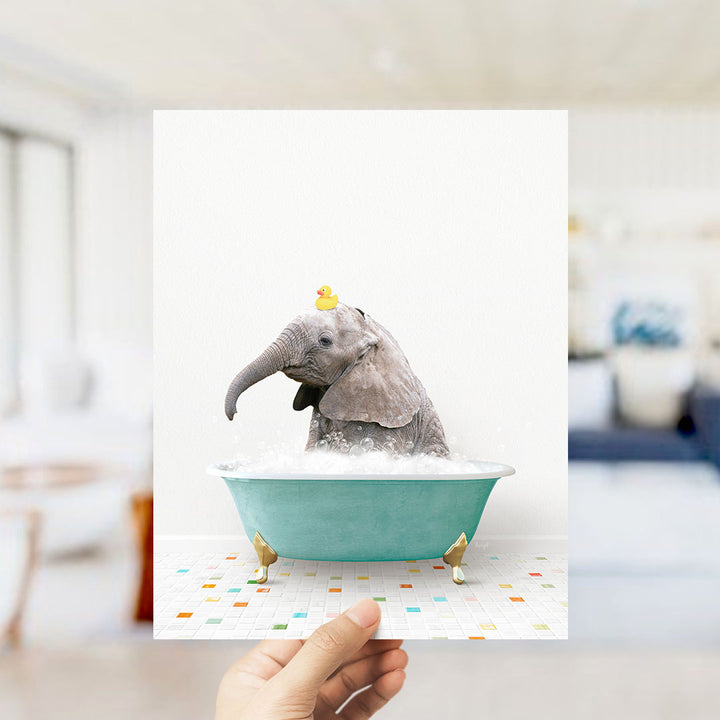 A hand holds up a framed image of a baby elephant sitting in a bathtub, with a yellow rubber duck floating on its head.
