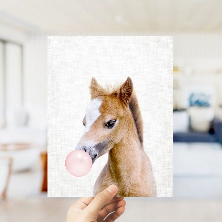 A hand is holding up a picture of a horse blowing a bubble with a pink bubble gum bubble.