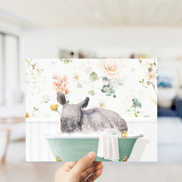 A hand holding a framed print featuring a rhinoceros sitting in a bathtub filled with bubbles, surrounded by a floral pattern.
