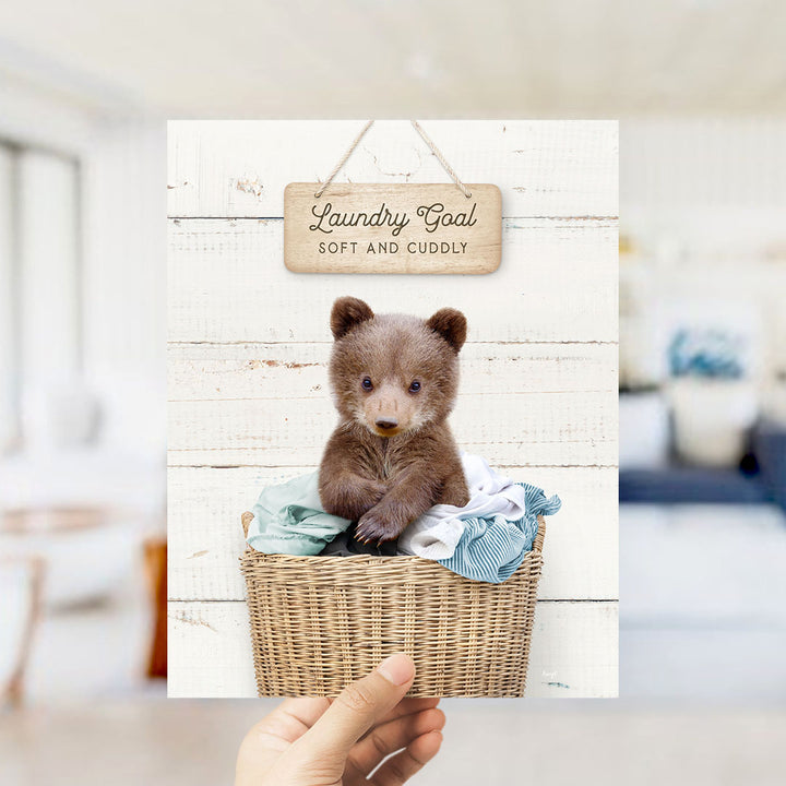 A hand holding a framed print with a cute brown bear sitting in a wicker basket filled with clothes, with a wooden sign above it that says "Laundry Goal Soft and Cuddly".