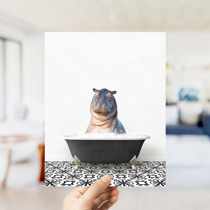 A hand holds up a framed image of a hippopotamus sitting in a bathtub filled with water, creating a whimsical and playful scene.
