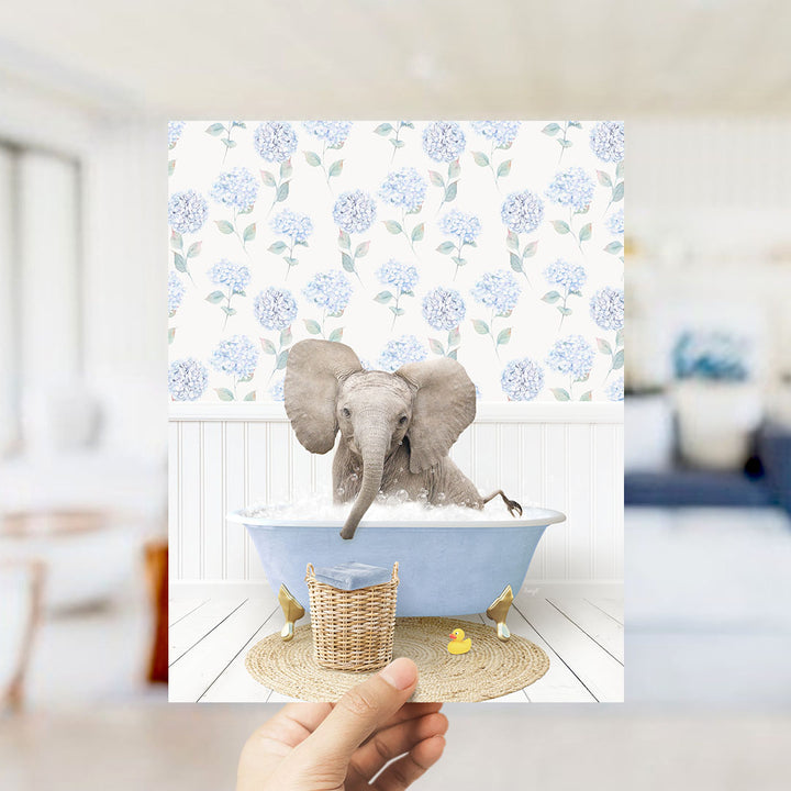 A hand holds up a framed image depicting a baby elephant sitting in a blue bathtub filled with water, surrounded by a blue and white floral pattern.