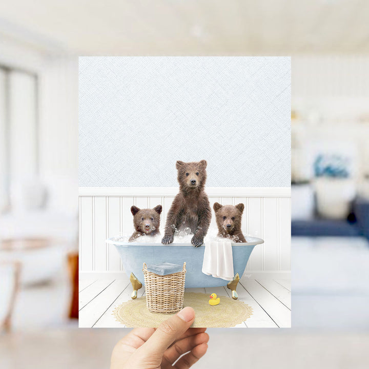 A hand holds up a framed image depicting three adorable brown bears sitting in a bathtub, with a yellow rubber duck floating in the water.