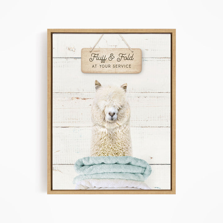 A framed image of a fluffy alpaca sitting on a stack of blue towels, with a wooden sign above it that says "Fluffy & Fold at your service".