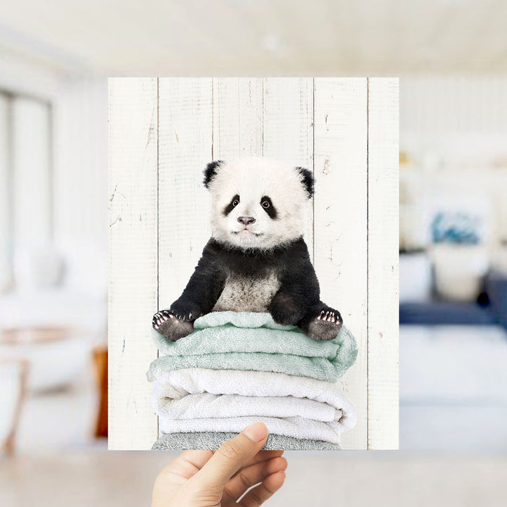 A hand is holding up a framed image of a panda bear sitting on a stack of towels.
