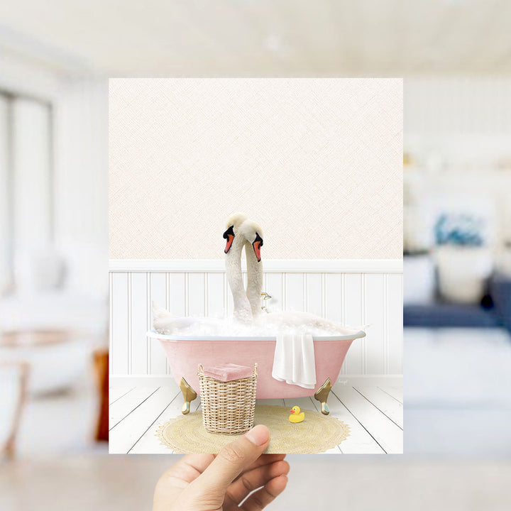 A hand holds up a framed image depicting two swans bathing in a pink bathtub, with a basket of rubber ducks nearby.