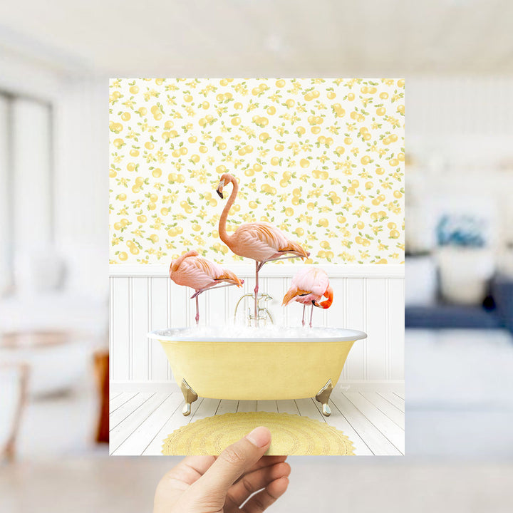 A hand holds up a framed image depicting three pink flamingos standing in a yellow bathtub filled with water, set against a yellow and white floral wallpaper.
