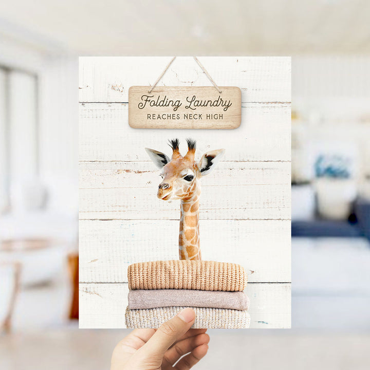 A hand is holding up a framed image of a giraffe standing on a pile of folded clothes, with a wooden sign above it that says "Folding Laundry Reaches Neck High".