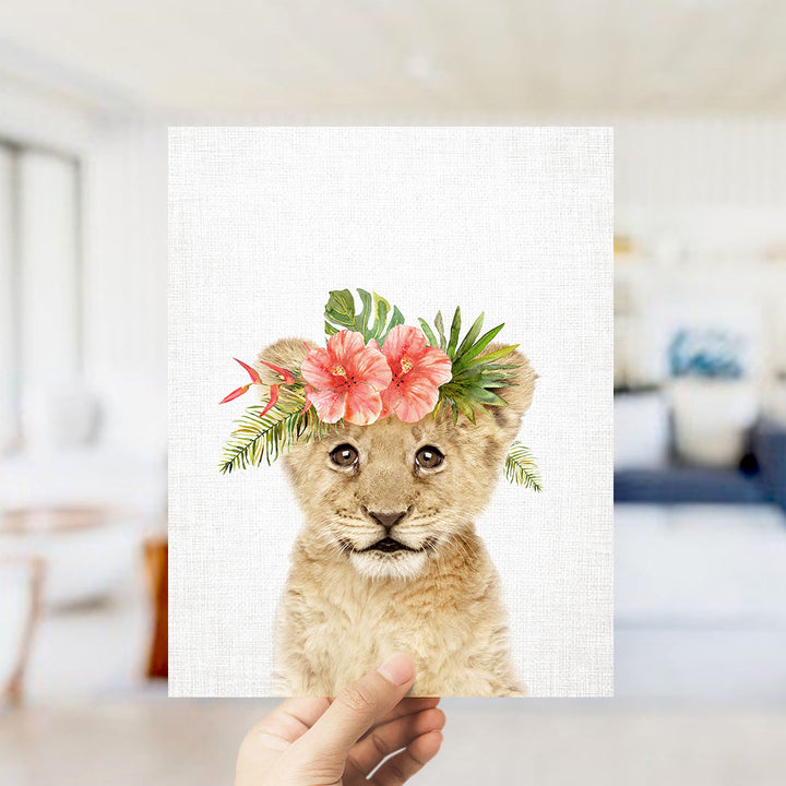 A hand is holding up a framed print of a young lion cub wearing a floral headpiece.