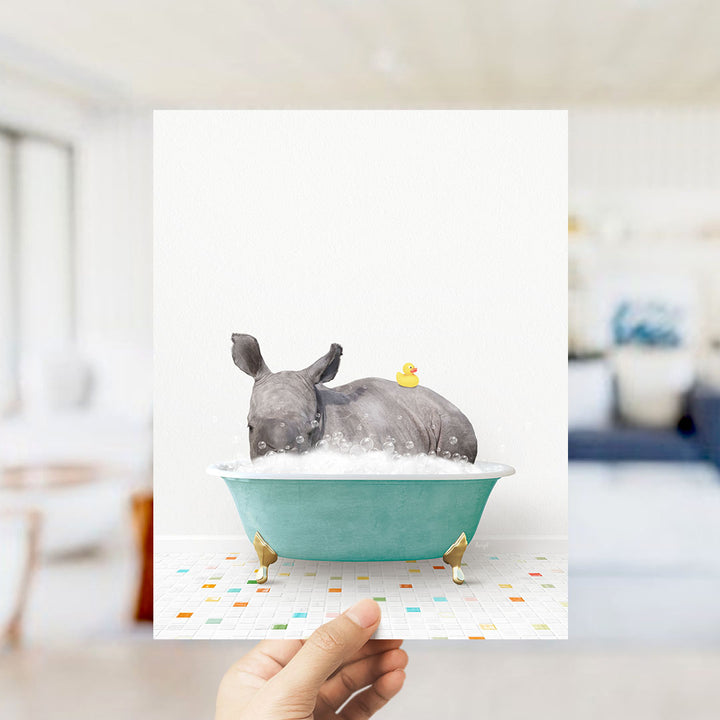 A hand holds up a framed image depicting a rhinoceros sitting in a teal-colored bathtub, with a yellow rubber duck floating on the water.
