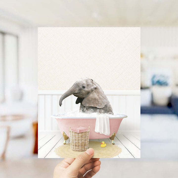 A hand holds up a framed image depicting a baby elephant sitting in a pink bathtub filled with bubbles, surrounded by a yellow rubber duck and a white towel.