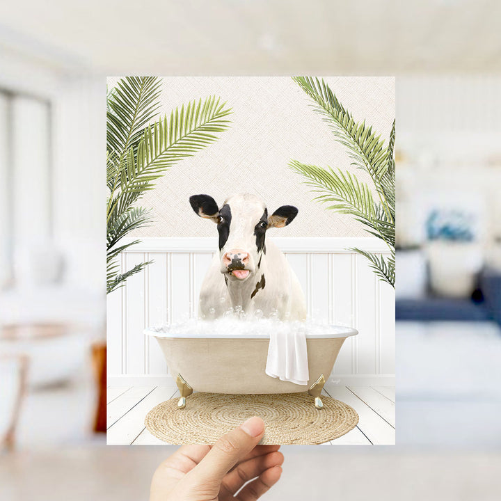 A hand holds up a framed image depicting a cow sitting in a bathtub filled with bubbles, surrounded by green palm leaves.