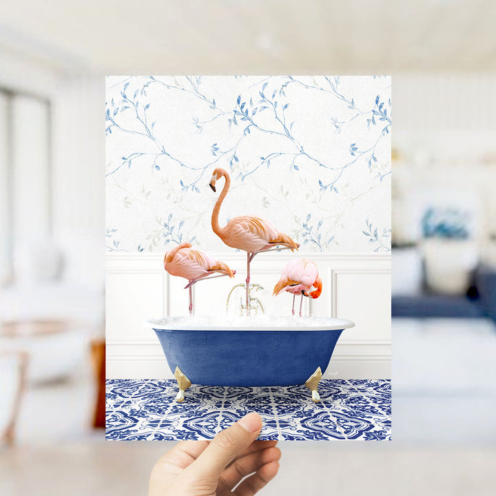 A hand holds up a framed image of three flamingos sitting in a blue bathtub against a floral wallpaper background.
