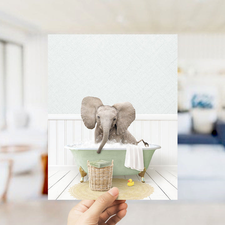 A hand holds up a framed image depicting a baby elephant sitting in a bathtub filled with water and bubbles, surrounded by a yellow rubber duck and a white towel.