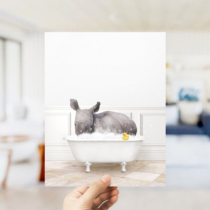 A hand holds up a framed image of a rhinoceros sitting in a bathtub filled with bubbles, with a yellow rubber duck floating in the water.