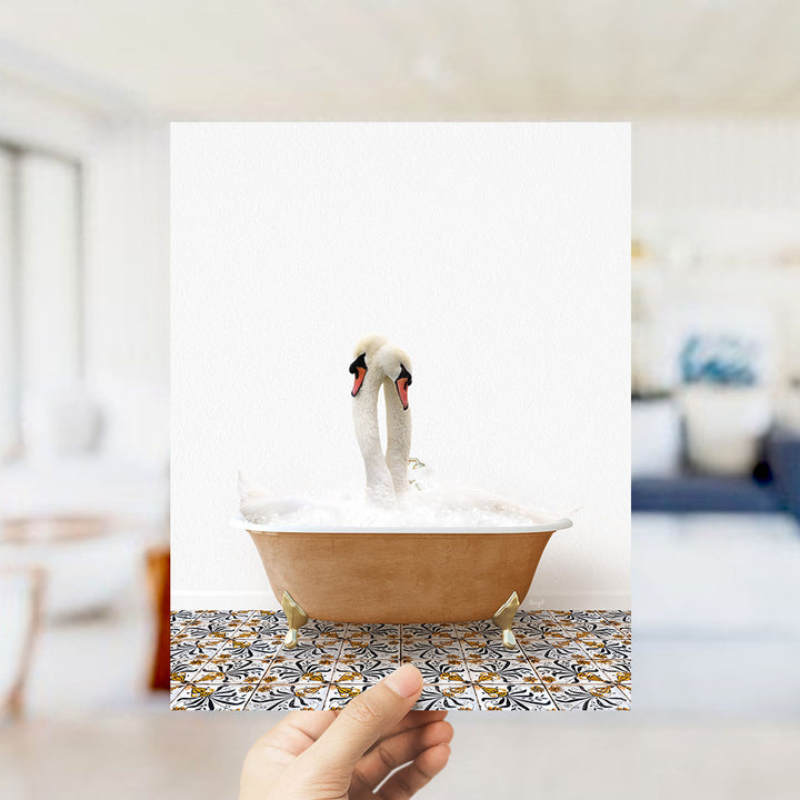 A hand holds up a framed photograph of two swans standing in a bathtub filled with bubbles.