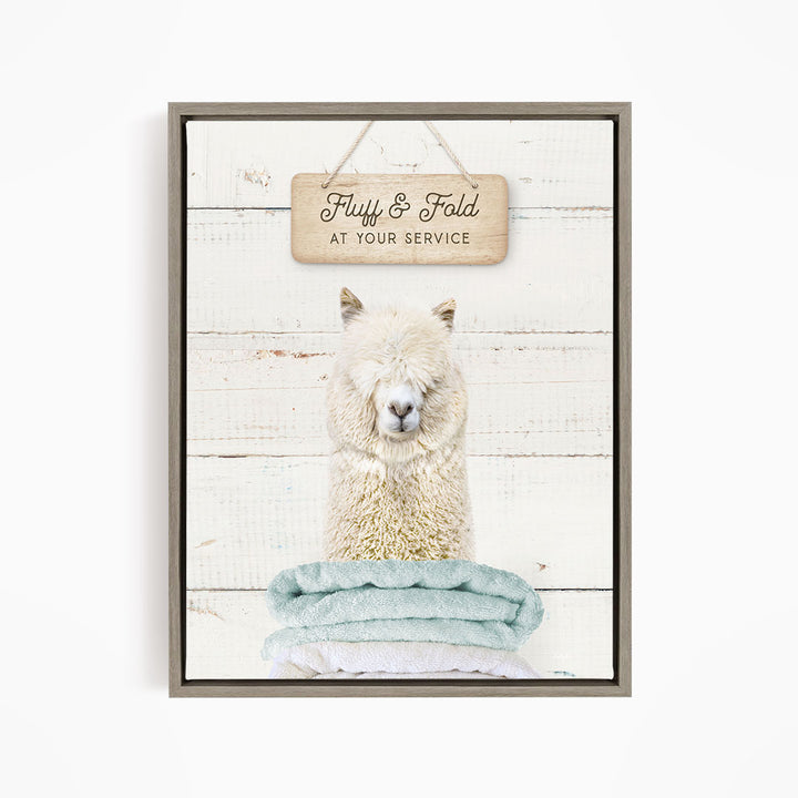 A framed image of a fluffy alpaca sitting on a blue towel, with a wooden sign above it that says "Fuffy & Fold at your service".