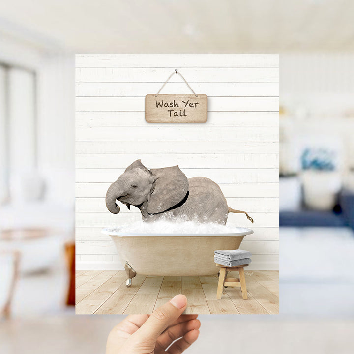 A hand holds up a framed image depicting a gray elephant sitting in a bathtub filled with water, with a sign above that reads "Wash Your Tail".