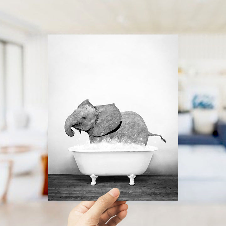 A hand holds up a black and white photograph of a baby elephant sitting in a bathtub filled with water.
