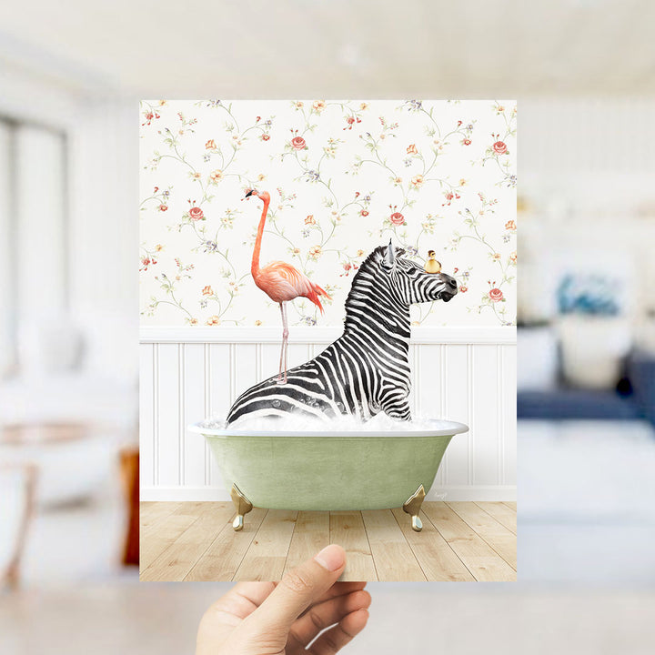 A hand holds up a framed image depicting a zebra sitting in a bathtub, with a pink flamingo standing on its back.