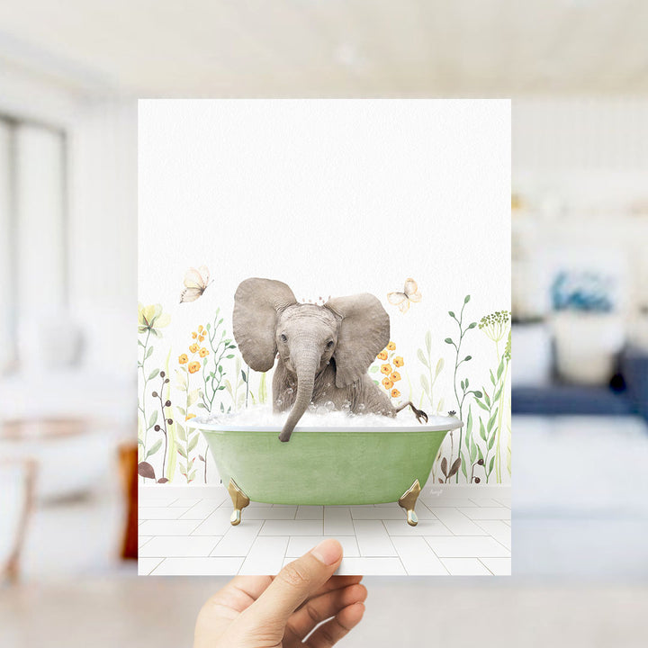 A hand holds up a framed image depicting a baby elephant sitting in a green bathtub filled with bubbles, surrounded by flowers and butterflies.