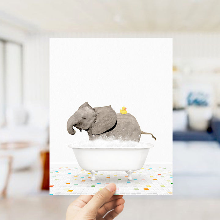 A hand holds up a framed image depicting a gray elephant sitting in a white bathtub, with a yellow rubber duck on its back.