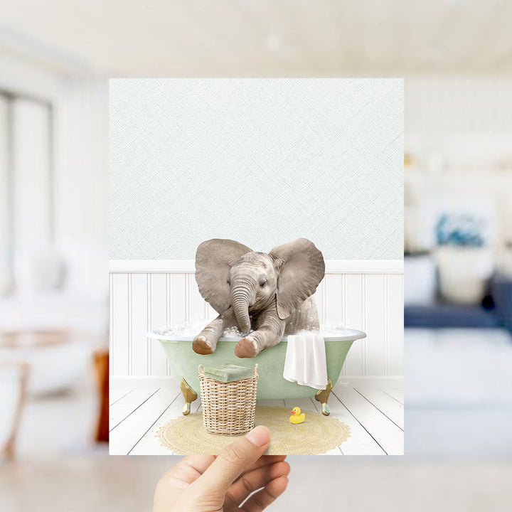 A hand holds up a framed image depicting a baby elephant sitting in a bathtub, surrounded by a basket of toys and a yellow rubber duck.