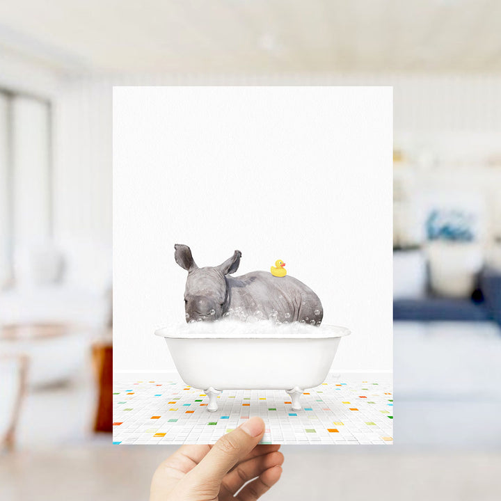 A hand holds up a framed image depicting a rhinoceros sitting in a bathtub filled with water, with a rubber duck floating on the water.