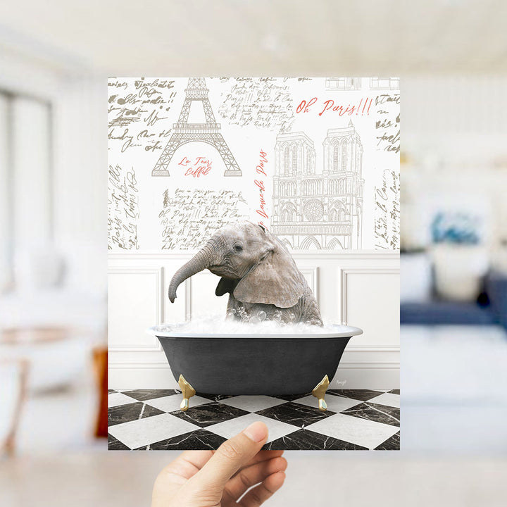 A hand is holding up a printed image featuring an elephant sitting in a bathtub filled with water, surrounded by various Paris-themed illustrations and text.