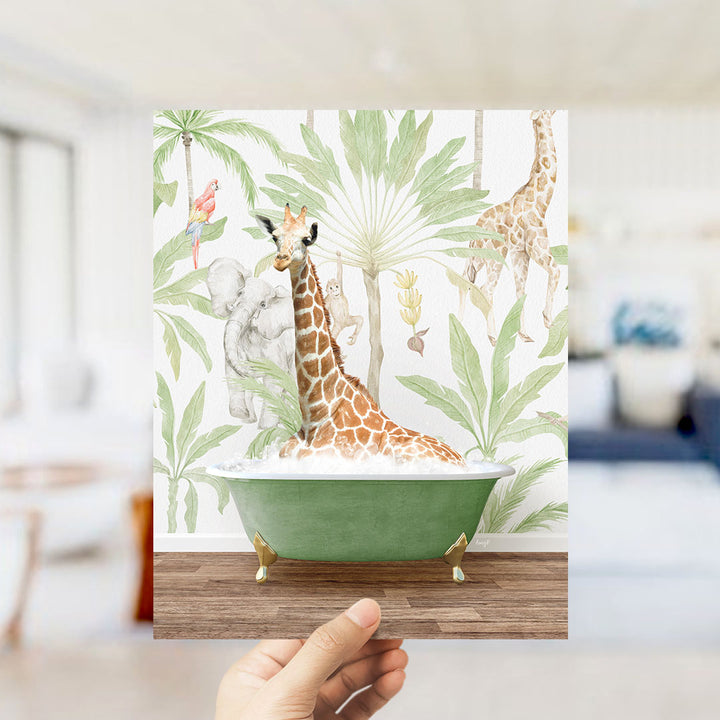 A hand holds up a framed print featuring a giraffe sitting in a green bathtub surrounded by tropical foliage.