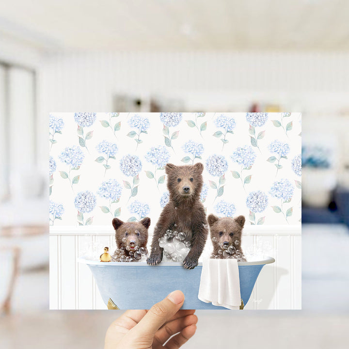 A hand holds up a collage of three images depicting three adorable brown bears sitting in a blue bathtub, surrounded by a floral pattern.