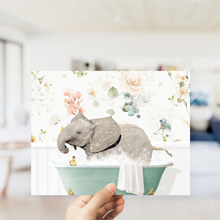 A hand is holding up a framed image depicting a gray elephant sitting in a green bathtub filled with water, surrounded by a floral pattern.