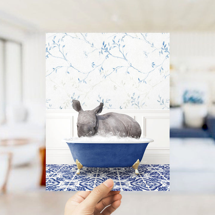 A hand is holding up a framed image of a rhinoceros sitting in a blue bathtub filled with bubbles, set against a blue and white floral wallpaper.